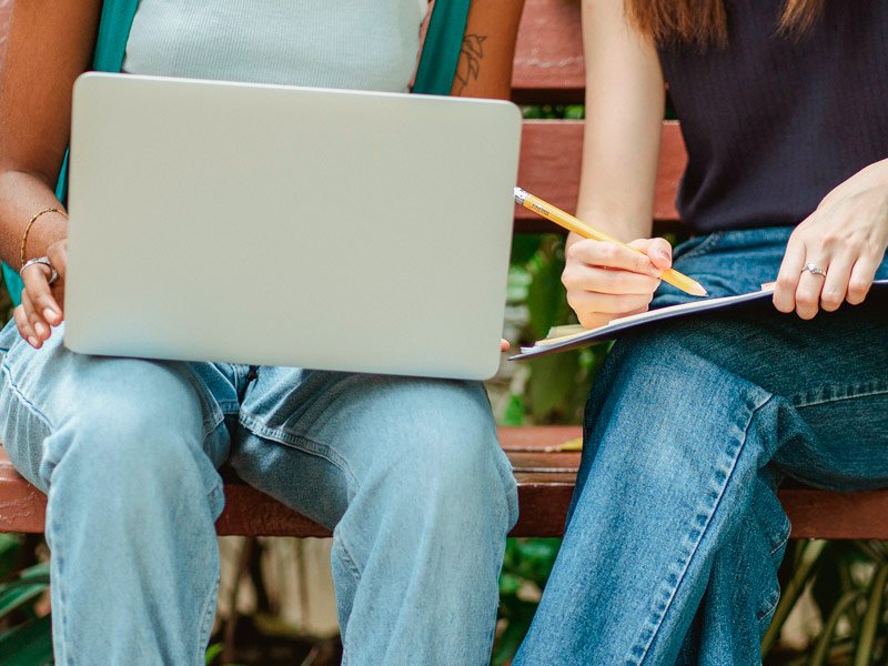 dos adolescentes estudiando con un cuaderno y un portátil sentadas en un banco en el exterior